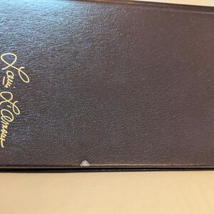 May include: A dark brown hardcover book with a textured finish. The front cover features the name Louis L'Amour embossed in gold script lettering. There are small areas of white wear visible along the bottom edge of the book cover.