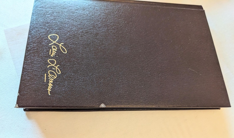 May include: A dark brown hardcover book with a textured finish. The front cover features the name Louis L'Amour embossed in gold script lettering. There are small areas of white wear visible along the bottom edge of the book cover.