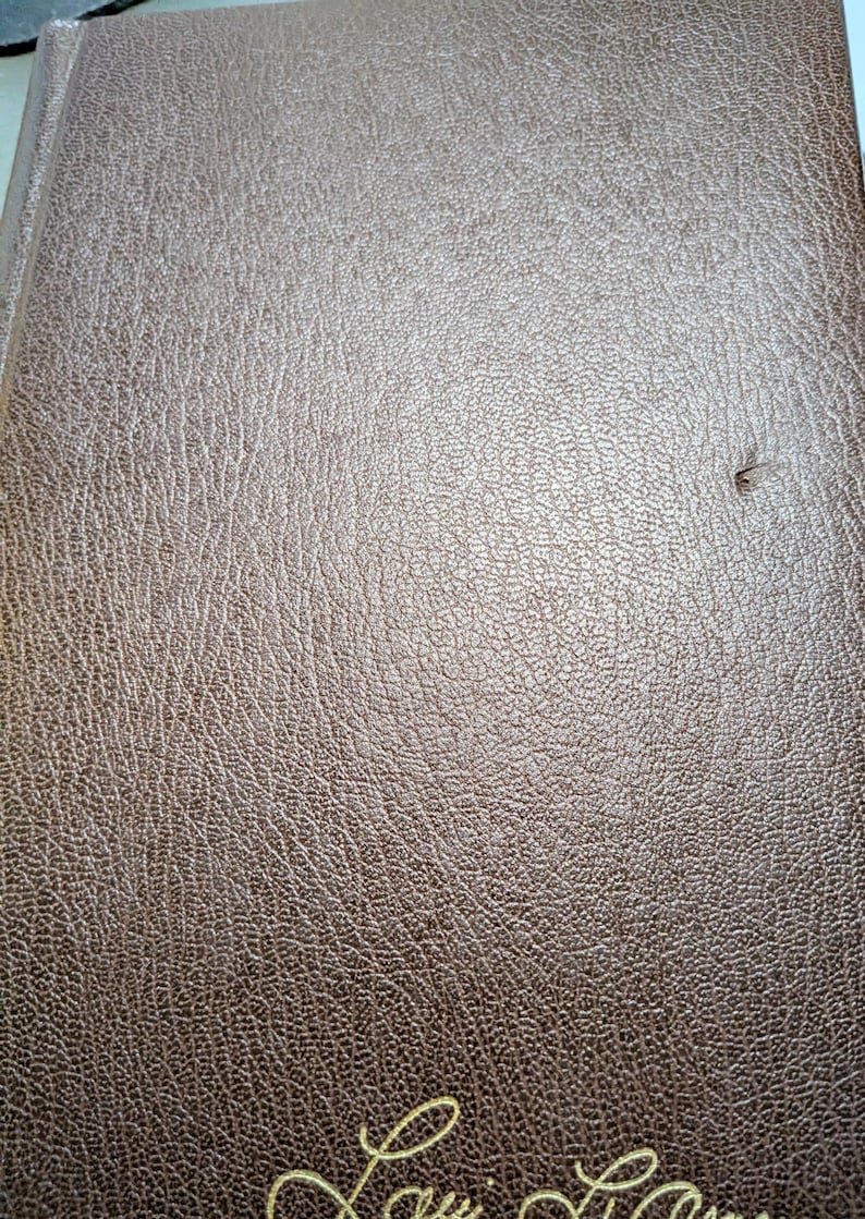 May include: A brown book with a textured, leather-like cover featuring a visible grain pattern. The bottom edge displays a partial, cursive gold-colored inscription. A small, dark indentation is present on the right side of the front cover.