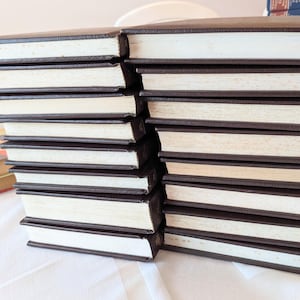 May include: A stack of fourteen hardcover books with dark brown covers and white pages arranged in two columns. The books are positioned horizontally, showing the page edges and the dark brown spines. A separate book with a yellow cover is visible.