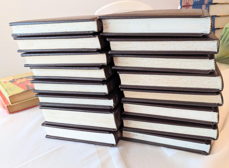 May include: A stack of fourteen hardcover books with dark brown covers and white pages arranged in two columns. The books are positioned horizontally, showing the page edges and the dark brown spines. A separate book with a yellow cover is visible.