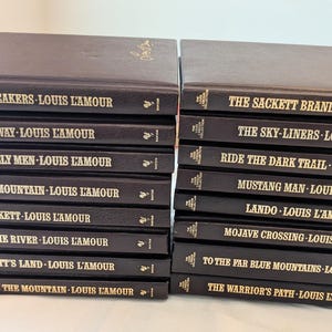 May include: A collection of sixteen hardcover books by Louis L'Amour, arranged in two stacks. Each book features a dark brown cover with gold lettering on the spine, displaying titles such as The Daybreakers, The Sackett Brand, and Galloway.