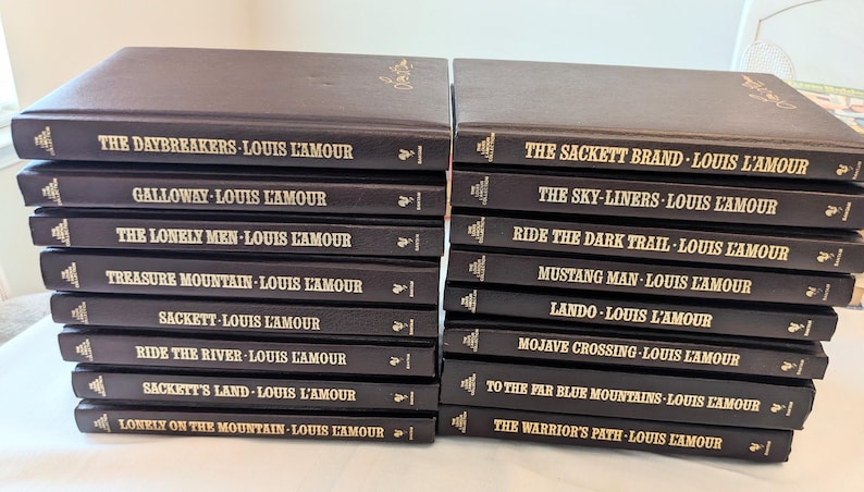 May include: A collection of sixteen hardcover books by Louis L'Amour, arranged in two stacks. Each book features a dark brown cover with gold lettering on the spine, displaying titles such as The Daybreakers, The Sackett Brand, and Galloway.