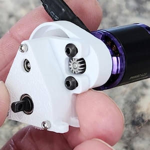 May include: A small, white, 3D-printed component with a black and purple motor. The component has a gear and is held by a person's fingers. The motor has a black body with purple accents. The component is secured with small screws.