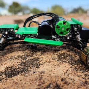 Puede incluir: Un chasis de vehículo a control remoto con marco negro, componentes impresos en 3D de color verde brillante y un motor eléctrico verde prominente. El conjunto incluye cuatro ruedas de metal dorado con radios intrincados y resortes visibles.