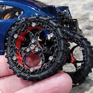 Puede incluir: Un coche de juguete azul oscuro con grandes neumáticos todoterreno negros personalizados con banda de rodadura texturizada. Las ruedas tienen un marco negro complejo y calado con detalles en rojo y herrajes plateados, fijado al lateral del vehículo.