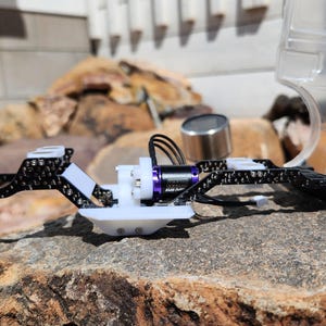 May include: A partially assembled small-scale model vehicle chassis, featuring a white and black frame, a purple motor, and a clear plastic body. The model is resting on a rock, with a blurred background.