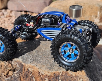 Puede incluir: Un coche teledirigido tipo rock crawler azul y negro con neumáticos grandes y con tacos y detalles azules. El vehículo está sobre una superficie rocosa, mostrando sus capacidades todoterreno. Un pequeño recipiente metálico es visible en el fondo.