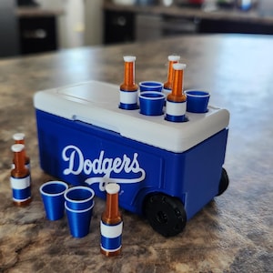 May include: A miniature blue and white cooler with the word "Dodgers" in white script. The cooler has a white lid, black wheels, and holds miniature bottles and blue cups. Several bottles and cups are placed around the cooler.