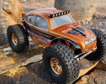 May include: A bright orange, custom-painted, off-road buggy with black accents and a black and white eagle graphic on the bonnet. The buggy has a black roll cage and a black and white sticker that reads "PunishmentRC x 2JORA RACING x ROVERIN". The buggy has a black and white sticker that reads "INJORA" on the side. The buggy has a light bar on the roof.