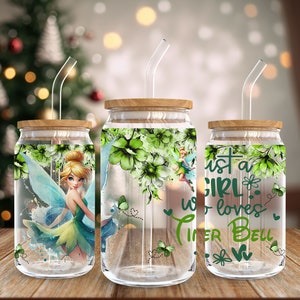 Puede incluir: Tres tarros de vidrio con tapas de madera y pajitas transparentes. Cada tarro tiene un diseño diferente. El primer tarro tiene un diseño floral verde con un personaje de Tinkerbell. El segundo tarro tiene un diseño floral verde. El tercer tarro tiene un diseño floral verde con el texto "Just a girl who loves Tinkerbell".