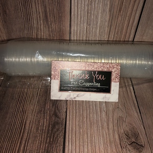 Puede incluir: Un paquete de plástico transparente que contiene una pila de vasos de plástico transparente. Una pequeña tarjeta con el texto "Thank You For Supporting" está delante del paquete.