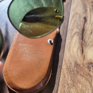 May include: Aviator-style sunglasses with gold frames and green lenses, resting on a brown leather case. The case is embossed with the Ray-Ban logo and the text "SUN GLASSES MADE IN U.S.A."