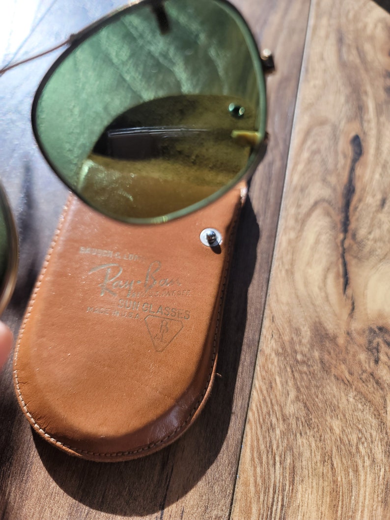 May include: Aviator-style sunglasses with gold frames and green lenses, resting on a brown leather case. The case is embossed with the Ray-Ban logo and the text "SUN GLASSES MADE IN U.S.A."