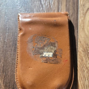 May include: A brown leather wallet with a curved bottom and a flap closure. The wallet has a faded design on the front and is stitched around the edges. The leather shows signs of wear.