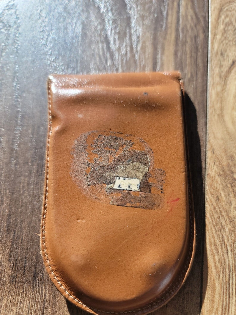 May include: A brown leather wallet with a curved bottom and a flap closure. The wallet has a faded design on the front and is stitched around the edges. The leather shows signs of wear.