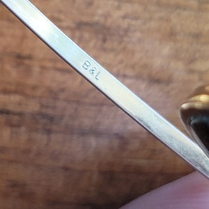 May include: Close-up of a silver metal object with the letters "B&L" engraved on it. The object is angled diagonally across the frame, with a blurred wooden background. The metal has a reflective surface.