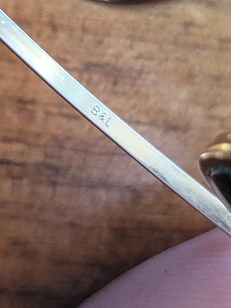 May include: Close-up of a silver metal object with the letters "B&L" engraved on it. The object is angled diagonally across the frame, with a blurred wooden background. The metal has a reflective surface.