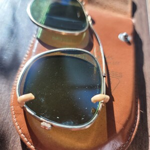May include: Vintage sunglasses with a gold frame and green lenses, resting in a brown leather case. The case is open, revealing the glasses. The sunglasses have a classic design, with a bridge and nose pads.