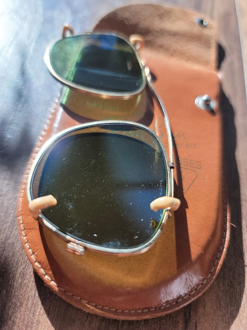 May include: Vintage sunglasses with a gold frame and green lenses, resting in a brown leather case. The case is open, revealing the glasses. The sunglasses have a classic design, with a bridge and nose pads.