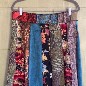 May include: A patchwork skirt with a variety of patterned fabrics in shades of red, teal, brown, and black. The skirt is hanging on a gray hanger against a light green background. The skirt's design includes floral, paisley, and abstract patterns.