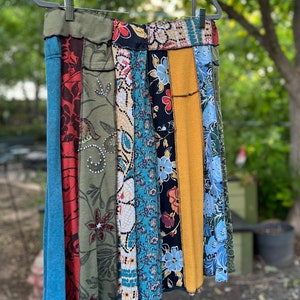 May include: A patchwork skirt with a variety of fabric patterns and colors, including blue, red, green, and yellow. The skirt is hanging on a black hanger against a blurred green background. The skirt has a unique, bohemian style.