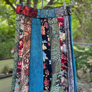 May include: A patchwork skirt with a variety of patterned fabrics in shades of red, green, blue, and brown. The skirt is hanging on a hanger against a blurred green background. The skirt's design includes floral, paisley, and abstract patterns.