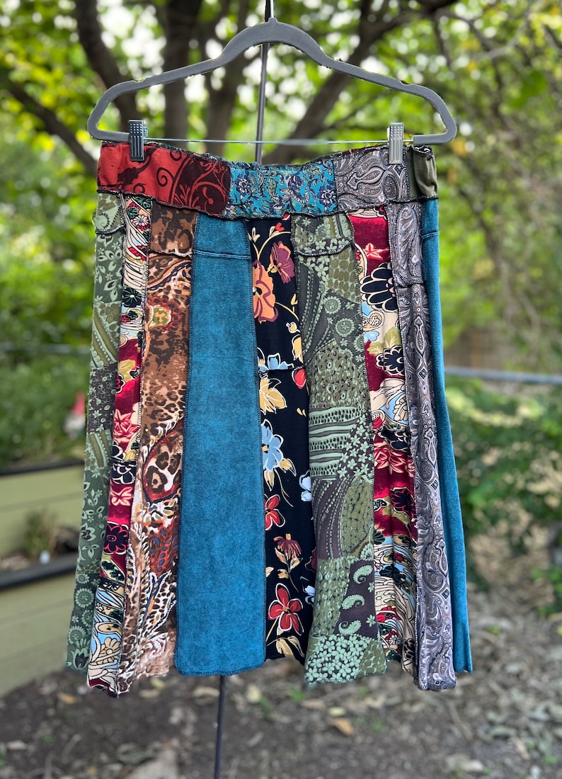 May include: A patchwork skirt with a variety of patterned fabrics in shades of red, green, blue, and brown. The skirt is hanging on a hanger against a blurred green background. The skirt's design includes floral, paisley, and abstract patterns.