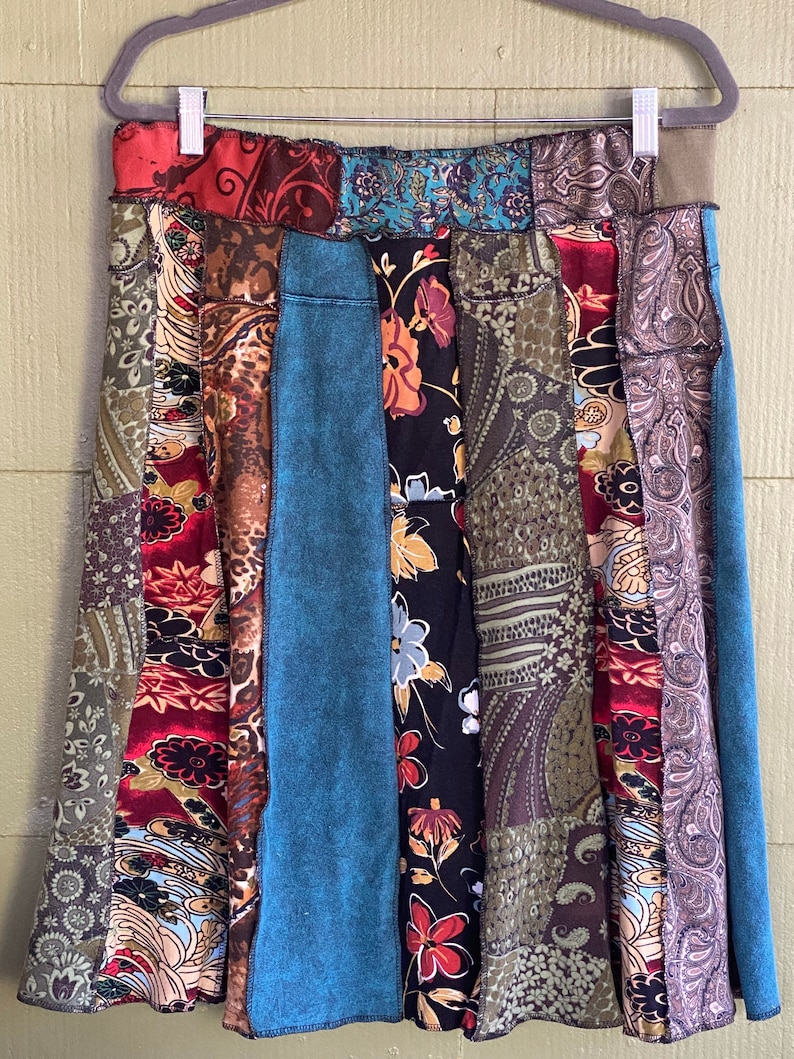 May include: A patchwork skirt with a variety of patterned fabrics in shades of red, teal, brown, and black. The skirt is hanging on a gray hanger against a light green background. The skirt's design includes floral, paisley, and abstract patterns.