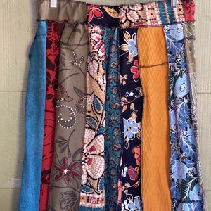 May include: A patchwork skirt with a variety of fabrics and patterns. The skirt features vertical panels of denim, floral prints, and textured fabrics in shades of blue, red, green, and gold. The skirt is hanging on a gray hanger.