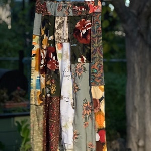 Könnte beinhalten: Ein Patchwork-Rock mit elastischem Bund, der an einem Kleiderbügel hängt. Der Rock hat verschiedene Stoffbahnen mit floralen und abstrakten Mustern in Grün-, Braun-, Rot- und Cremetönen. Der Rock ist knielang.