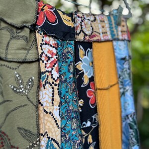 May include: A patchwork skirt with a variety of fabric patterns and colors, including olive green, black with red flowers, teal floral, and mustard yellow corduroy. The skirt is hanging on a metal hanger outdoors.