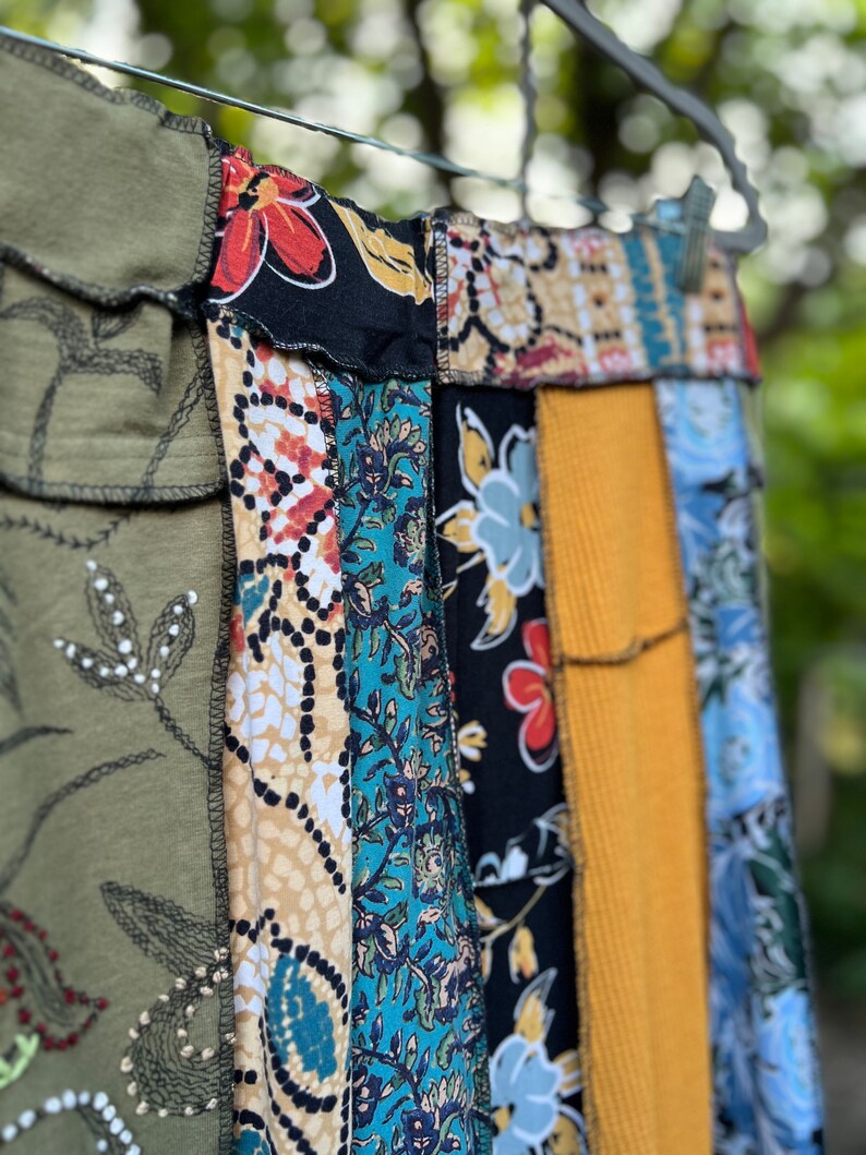May include: A patchwork skirt with a variety of fabric patterns and colors, including olive green, black with red flowers, teal floral, and mustard yellow corduroy. The skirt is hanging on a metal hanger outdoors.