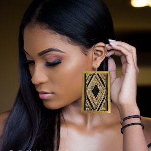 May include: A pair of large, gold-toned earrings with a black geometric design. The earrings are rectangular in shape and feature a diamond-shaped pattern with black accents. The earrings are worn by a person with long black hair.