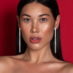 以下が含まれることがあります： 滑らかで光沢のある仕上げが施された、長く細いシルバー調のバー型ピアス。耳たぶから垂直にぶら下がるデザインで、顔の横に沿ってまっすぐに伸びるシンプルで直線的な形状が特徴です。
