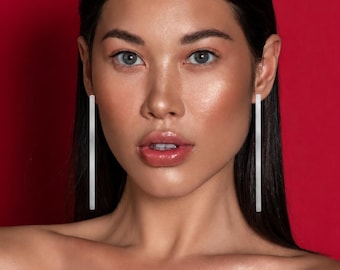 May include: A pair of long, slender, silver-toned bar earrings with a smooth, polished finish. The earrings hang vertically from the earlobes, featuring a clean, straight-line design that extends downward along the sides of the face.