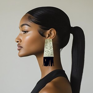 May include: A side profile of a person wearing a black top and large, rectangular earrings. The earrings are cream-colored with a geometric pattern and black fringe.
