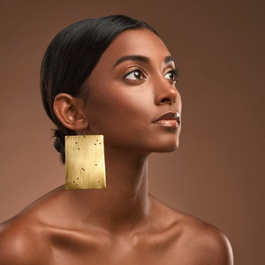 May include: A woman with dark hair and brown skin is wearing large gold rectangular earrings with a textured surface. She is looking up and to the right.