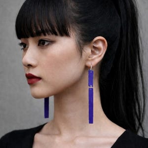 May include: A person with black hair and a ponytail wears long, rectangular, cobalt blue earrings. The earrings are suspended from small silver hooks. The person is wearing a black top and has dark eye makeup and red lipstick.