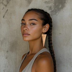 Könnte beinhalten: Eine Frau mit geflochtenem dunklem Haar trägt ein beiges Top und große, goldene, dreieckige Ohrringe. Die Ohrringe hängen an einem dunklen Metallhaken. Die Frau schaut in die Kamera.