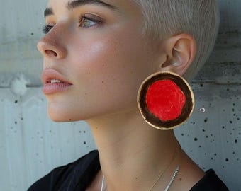 Puede incluir: Un pendiente circular grande que presenta un centro rojo vibrante rodeado por un borde marrón oscuro. El pendiente se lleva en el lóbulo de la oreja y tiene una forma de disco plano con un acabado liso, contrastando con la ropa oscura y el pelo claro.