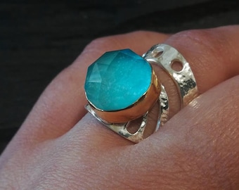 Puede incluir: Un anillo con una gran piedra redonda facetada de color turquesa engastada en un bisel dorado. La banda está hecha de metal plateado martillado y presenta una serie de formas circulares recortadas a lo largo de la superficie ancha y texturizada.