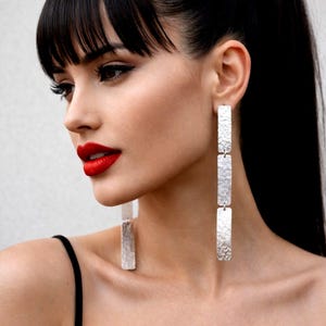 May include: Silver rectangular drop earrings with a hammered texture. The earrings are long and dangle, with three connected sections. The model has dark hair and red lipstick, wearing a black top.