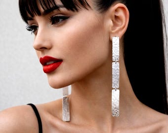 Peut inclure: Boucles d'oreilles pendantes rectangulaires en argent avec une texture martelée. Les boucles d'oreilles sont longues et pendantes, avec trois sections connectées. Le modèle a les cheveux foncés et du rouge à lèvres rouge, portant un haut noir.