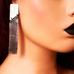 Puede incluir: Un par de pendientes de plata con un diseño rectangular texturizado. Los pendientes son largos y cuelgan del lóbulo de la oreja.
