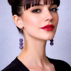 Op de afbeelding: Close-up van een persoon die lange, bungelende oorbellen draagt. Elke oorbel heeft drie ronde, getextureerde, paarse kralen die aan een dunne, zilverkleurige staaf hangen. De persoon draagt een zwarte top en heeft donker haar en rode lippenstift.