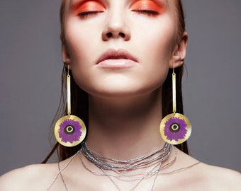 May include: A woman with red hair and orange eyeshadow wears long gold earrings with purple flower accents. The earrings are dangling and have a round shape.