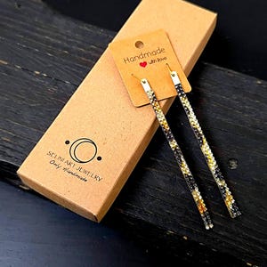 Può includere: Orecchini pendenti lunghi con ganci color oro e un design lineare di cristalli neri, dorati e trasparenti. Gli orecchini sono esposti su un cartoncino e una scatola marrone. L'etichetta recita "Handmade with love".