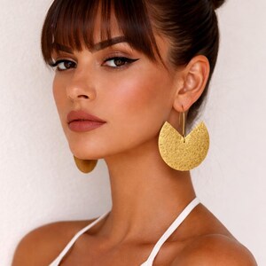 May include: Gold-colored, textured half-circle earrings. The earrings are suspended from a thin gold wire. The model is wearing a white top and has dark hair pulled back. The earrings are a statement piece.