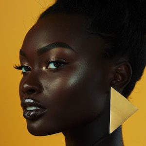 Op de afbeelding: Een vrouw met een donkere huid en donker haar in een knot. Ze draagt grote, gouden, driehoekige oorbellen. De achtergrond is warmgeel. Haar lippen zijn donker en haar ogen zijn opgemaakt. De afbeelding is een close-up.